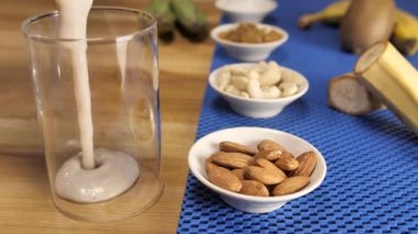 Çeşit çeşit kuru meyveli olgun muzlu milkshake şeffaf bir bardağa, sağlıklı protein karışımına dökülüyor. Yakın çekim badem, kaju, kuru üzüm, muz, kivi ve şeker mavi masa paspası üzerinde bir arada tutulur.