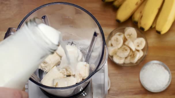 健康的なスムージーを作るためにバナナのスライスとブレンダーに新鮮な牛乳を注ぐ 夏の飲み物インド 黄色の熟したバナナとバナナの部分 砂糖のボウル 木製のテーブルの上にミルクシェイクを置くミキサー — ストック動画