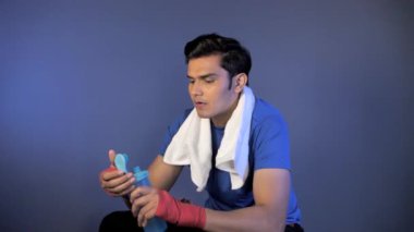 Eldivenleri ve havlusu olan bir sporcu, koyu bir arka planda oturarak, bir yudumdan sağlık içeceği içiyor. Genç Hintli adam günlük antrenmanından sonra dinlenirken su içiyor - sağlıklı aktif yaşam tarzı