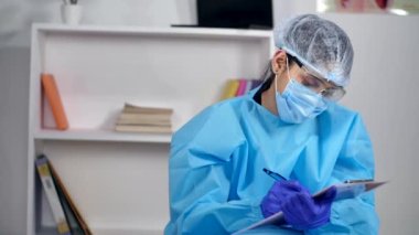 Tıbbi koruyucu giysili bir sağlık görevlisi bir not panosuna not alıyor. Küresel salgın sırasında PPE Kit ile hastaların raporlarını yazan bir hastane personelinin orta boy fotoğrafı.