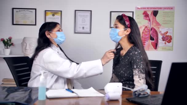 Covid 19症状のための若い患者を調べるために聴診器を使用している医師 白人の制服を着たインド人女性医師と彼女の患者を診断する医療マスク 医療サービス — ストック動画
