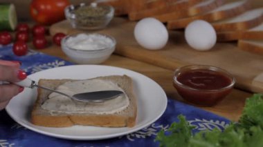 Bir kaşık mayonezi bir dilim kahverengi ekmekli sandviçe süren kadın elleri. Yakın plan iki yumurta, domates ketçap, marul yaprakları ve ahşap bir masada bir arada tutulan beyaz ekmek dilimleri.