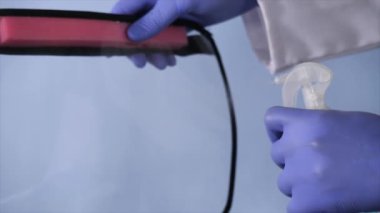 A medical worker in a white coat disinfecting the face shield using a sanitizer. Closeup shot of a male doctor with blue gloves taking precautions wearing a face shield before examining a patient