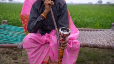 Yaşlı bir bayanın boş zamanlarını Hint piposu içerek geçirdiği orta boy bir fotoğraf. Yeşil tarım arazisinin yakınındaki örülmüş bir karyolada oturan yaşlı bir kadın dinleniyor ve nargile yiyor.