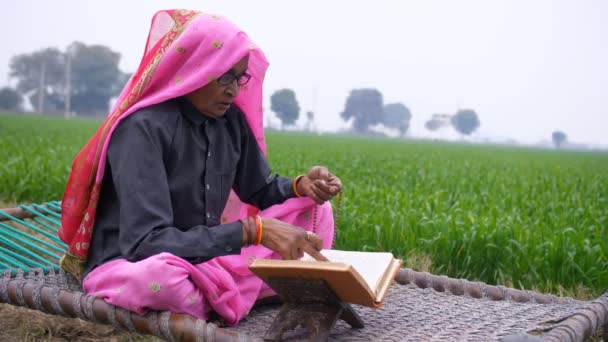 宗教書を読んで神を崇拝する村の老女 インド人の老婦人が神聖な本を読み 木のビーズを使って神の名を唱えながら宗教的信念に従って — ストック動画