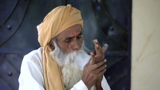 額にオレンジのティカを持ったインド人の老人がタバコを吸っていた 髭を生やした農村の村人は 自宅で座ってリラックスしながら喫煙を楽しんでいます ライフスタイルと中毒 — ストック動画