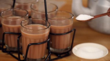 İnsan eli, bir bardak çaya bir kaşık şeker koyuyor. Bardak tutacağında saklanıyor. Hint çayı. Yakın planda paslanmaz çelik bir su ısıtıcısı ve bir kase şeker, ahşap bir masada serinletici bir içecek olarak duruyordu.