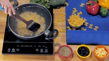 Ahşap bir spatulayla çubuksuz tencerede farfalle makarna karıştıran bir kadın. Suyun içinde kaynamış buğday makarnası ve ahşap bir masaya yerleştirilmiş taze sebzeler.