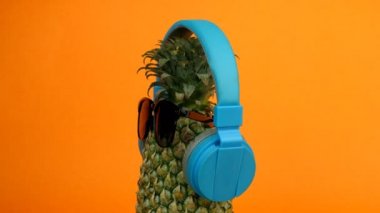 Gözlüklü ve kulaklıklı ananas turuncu arka planda yavaşça dönüyor. Tropikal yaz tatili kavramı - Olgun ve sulu Ananas sağlıklı antioksidanlar, güneş gözlüğü ve kulaklık ile zengindir