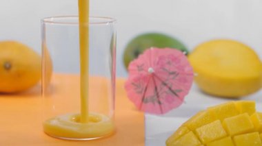 Serinletici mango milkshake ve Lassi 'nin üst görüntüsü berrak bir bardağa döküldü - Hindistan Yazı İçeceği. Yakın çekim sarı mango, yeşil mango, kokteyl şemsiyesi ve mango dilimleri ahşap bir masada bir arada tutulur.