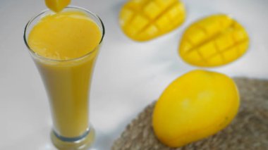 Bir bardak taze mango milkshake 'ine düşen küçük küpler. Sağlıklı içecek, Hindistan Yazı içeceği. Tropikal meyvelerin yakın plan çekimi lezzetli Aam dilimleri ve yarımları ahşap bir masada bir arada tutulur.
