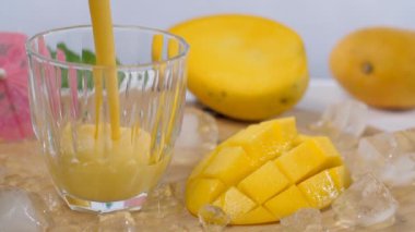 Şeffaf bir bardağa dökülen taze mango suyu / smoothie - Hindistan yazı içeceği. Yakın çekim sarı mango dilimler halinde kesilmiş ve birkaç buz küpü ahşap bir masada bir arada tutulmuş.