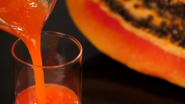 Sabah kahvaltısı için şeffaf bardağa tropik papaya meyve suyu döküldü. Siyah arka plana yerleştirilmiş tohumlarla ikiye bölünmüş turuncu bir papaya sağlıklı beslenme