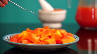 Kadının elleri seramik tabaktan çatalla sulu bir papaya alıyor. Kavanozda turuncu papaya ezmesi ve tropikal meyve suyu var. Mavi arka planda tutuluyor. Sağlıklı beslenme.