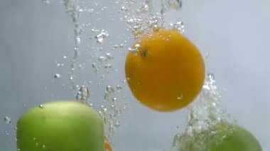 Tüm yeşil elmalar ve şeffaf suya dökülen soyulmamış sarı portakallar. Suda beyaz bir zemin üzerinde yüzen sağlıklı ve besleyici meyvelerin yakın plan çekimi