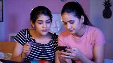 Neşeli kız en iyi arkadaşına online alışverişte yardım ediyor. E-ticaret alışverişinde. Genç Hintli kız kardeşler yatak odasında birlikte oturuyorlar. Cep telefonlarıyla alışveriş yapıyorlar. İnternetten alışveriş yapıyorlar.
