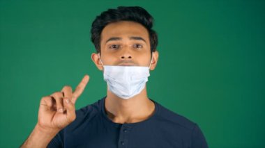 Yakışıklı genç adam Coronavirus sırasında tıbbi maskeyi çıkarmaktan kaçınıyor. Çekici bir adam kameraya doğru başparmak hareketleriyle bakıyor. Poz veriyor, modern yaşam tarzı, sosyal mesafeler, Covid-19.