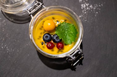 Cam kavanozda taze tatlı, tutku meyvesi ve böğürtlenli. Sarı tutku meyveli jöle soslu panna cotta siyah taş kapaklı cam bir tencerede servis edilir.