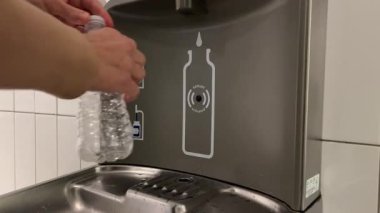 Birisi su deposunda plastik bir su şişesini dolduruyor. Mekan temiz ve parlak, modern özellikler ve açık işaretler gösteriyor..