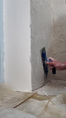 Bir insan köşedeki duvarda alçıyı düzleştiriyor. İş kapalı alanda iyi ışıklandırmayla olur. Görev için araçlar ve malzemeler düzenlenmiştir.