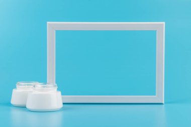 Beyaz cilt kremi kavanozu ve mavi arka planda beyaz fotoğraf çerçevesi. Mesaj için yer. Boşluğu kopyala