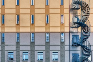 Gri, sarmal bir merdiveni ve dikdörtgen, küçük bir pencere deseni olan bir binanın ön cephesinde minimal endüstriyel mimari. Gri yangın merdivenlerinin ön görüntüsü veya pembe çıplak modern dış yüzeydeki küçük dikdörtgen pencereli merdiven.