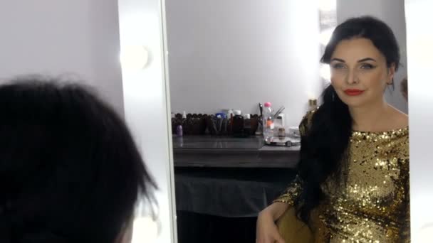 A beautiful spectacular young woman with a stunning evening make up smoky eyes in a sparkling gold dress with sequins sits in front of a mirror and looks at her reflection — Stock Video