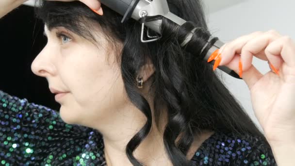 A beautiful spectacular young woman winds her long black hair on a special iron to create curls close up view — Stock Video