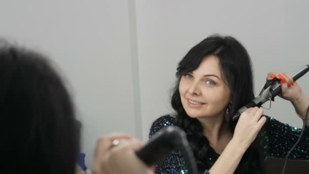 A beautiful spectacular young woman in front of a mirror winds her long black hair on a special iron to create curls — Stock Video