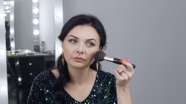 Joven hermosa mujer vistosa delante de un espejo aplica maquillaje y fundación o corrector con un cepillo especial — Vídeos de Stock