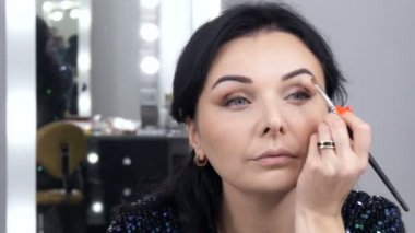 Aynanın önündeki güzel, gösterişli genç kadın özel bir fırçayla yüz ve göz makyajı yapıyor.