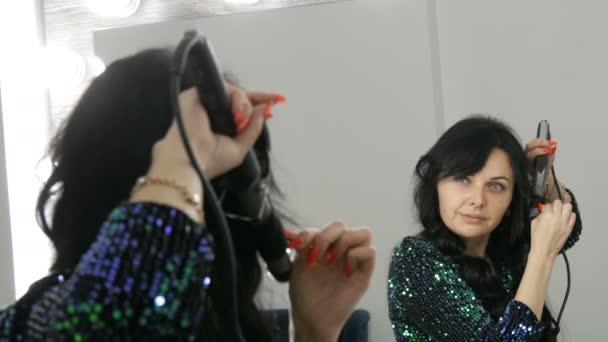 A beautiful spectacular young woman in front of a mirror winds her long black hair on a special iron to create curls — Stock Video