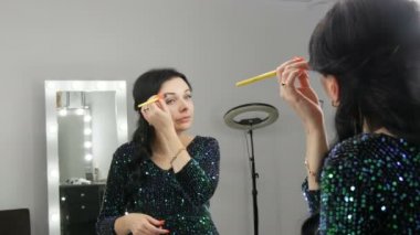 Aynanın önündeki güzel, gösterişli genç kadın özel bir fırçayla yüz ve göz makyajı yapıyor.