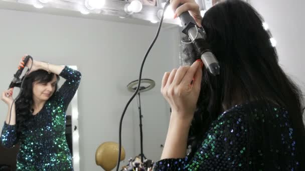 Una hermosa joven espectacular delante de un espejo enrolla su largo cabello negro sobre una plancha especial para crear rizos — Vídeos de Stock