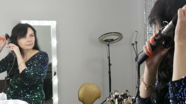 Aynanın önünde muhteşem bir genç kadın uzun siyah saçlarını bukleler yaratmak için özel bir ütüye sarıyor. — Stok video