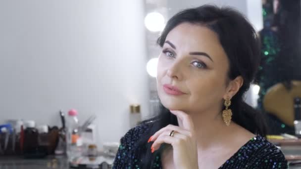 Uma bela jovem vistosa com uma deslumbrante noite make-up olhos fumegantes em um vestido azul espumante com lantejoulas senta-se na frente de um espelho e olha para o seu reflexo — Vídeo de Stock