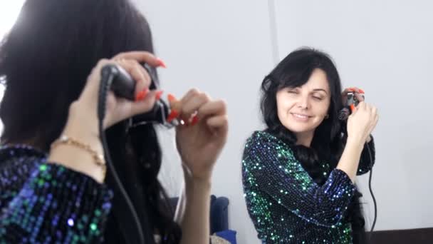 A beautiful spectacular young woman in front of a mirror winds her long black hair on a special iron to create curls — Stock Video