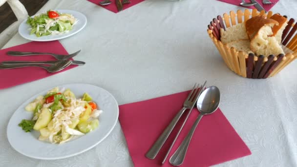 Table setting in the restaurant. Bread in a basket, vegetable salad in a plate, cutlery knife, fork, spoon on a red napkin top view — Stock Video