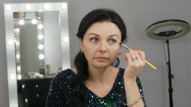 Aynanın önündeki güzel, gösterişli genç kadın özel bir fırçayla yüz ve göz makyajı yapıyor.