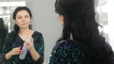 Genç ve güzel bir kadın aynanın önünde saçını yapıyor. Uzun siyah saçlı. Saçına bir şişe saç spreyi sıkılmış.