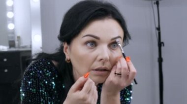 Aynanın önündeki güzel, gösterişli genç kadın özel bir fırçayla yüz ve göz makyajı yapıyor.