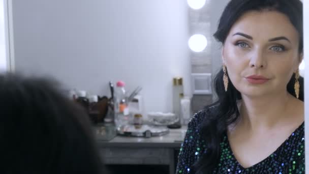 A beautiful showy young woman with a stunning evening make-up smoky eyes in a sparkling blue dress with sequins sits in front of a mirror and looks at her reflection — Stock Video