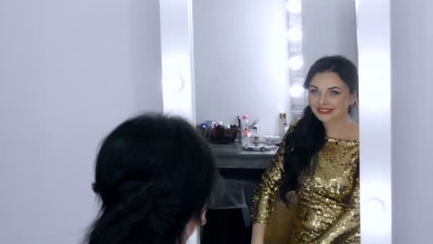 Een mooie opzichtige jonge vrouw met een prachtige avond make-up rokerige ogen in een sprankelende gouden jurk met pailletten zit voor een spiegel en kijkt naar haar reflectie — Stockvideo