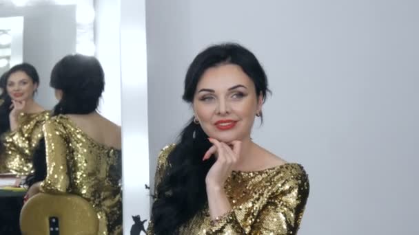 Een mooie opzichtige jonge vrouw met een prachtige avond make-up rokerige ogen in een sprankelende gouden jurk met pailletten zit voor een spiegel en kijkt naar haar reflectie — Stockvideo