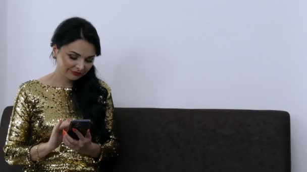 Krásná mladá žena s večerní make-up v krásných večerních zlatých šatech s flitry tiskne něco a mluvit úsměv na telefonu v místnosti — Stock video