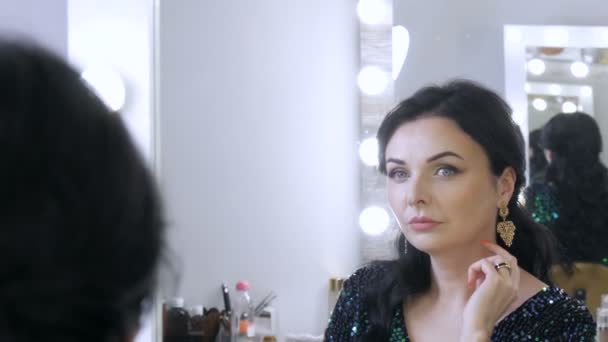 Uma bela jovem vistosa com uma deslumbrante noite make-up olhos fumegantes em um vestido azul espumante com lantejoulas senta-se na frente de um espelho e olha para o seu reflexo — Vídeo de Stock