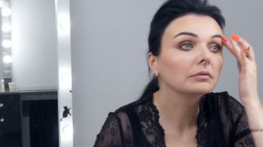 Siyah bir peignoir ve yakası olan güzel bir genç kadın aynanın önünde özel bir fırçayla yüzüne makyaj yapıyor.