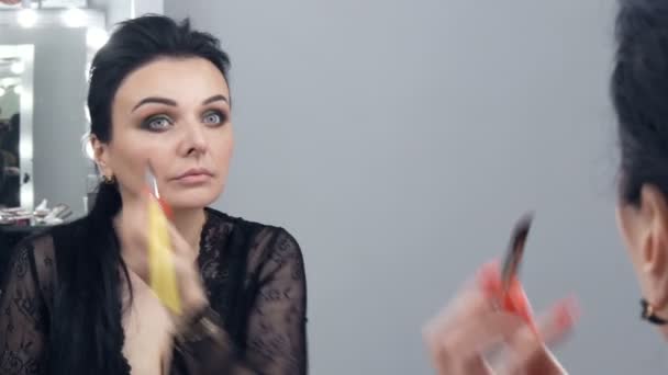 Seorang wanita muda yang spektakuler dalam sebuah peignoir hitam dan kalung menerapkan makeup ke wajahnya dengan kuas khusus di depan cermin — Stok Video
