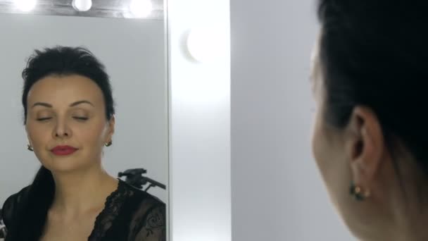 Frumoasă femeie tânără cu păr lung și într-un peignoir negru și decolteu se uită la reflecția ei în oglindă . — Videoclip de stoc