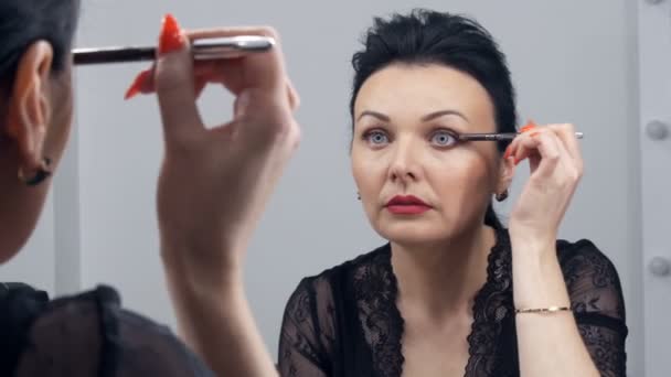 Uma bela jovem espetacular em um peignoir preto e decote aplica maquiagem em seu rosto com uma escova especial na frente de um espelho — Vídeo de Stock